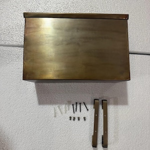 May include: A rectangular, antique brass-finished metal mailbox with a flat top lid. Included are two vertical mounting brackets, four black screws, four small metal fasteners, and four plastic wall anchors for installation purposes.