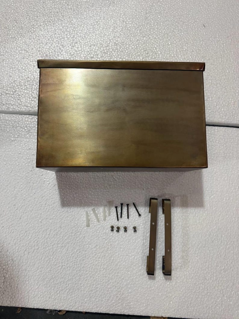 May include: A rectangular, antique brass-finished metal mailbox with a flat top lid. Included are two vertical mounting brackets, four black screws, four small metal fasteners, and four plastic wall anchors for installation purposes.