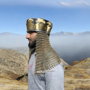 May include: A gold and silver helmet with a chainmail coif. The helmet has a crown-like design and is worn by a person standing on a rocky mountaintop. The text "Handmadelightinghub" is visible on the chainmail.