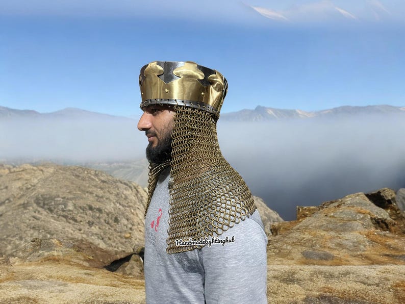 May include: A gold and silver helmet with a chainmail coif. The helmet has a crown-like design and is worn by a person standing on a rocky mountaintop. The text "Handmadelightinghub" is visible on the chainmail.
