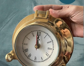May include: A 6 inch brass clock with Roman numeral markings on a white face. The clock is in a round, brass case with a clear glass cover. The clock is being held in a hand.