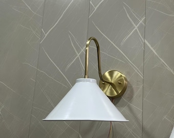 May include: A white, cone-shaped wall sconce with a brass-coloured curved arm and circular base. The sconce is mounted on a grey wall with white veining. The fixture has a minimalist design.