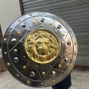 May include: A circular shield in silver with a gold lion's head emblem at its centre. The shield features a decorative border with rivets and circular accents. The lion's head is intricately detailed with a mane and facial features.