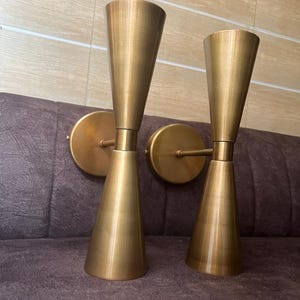 Op de afbeelding: Twee messingkleurige wandkandelaars met een modern design uit het midden van de eeuw. Elke wandkandelaar heeft een conische kap en een cirkelvormige wandbevestiging. De wandkandelaars zijn gemaakt van metaal en hebben een warme, gouden afwerking.