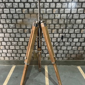 Puede incluir: Una l&aacute;mpara de pie de madera con un tr&iacute;pode de metal plateado y una pantalla negra. La l&aacute;mpara est&aacute; sobre un suelo de baldosas.