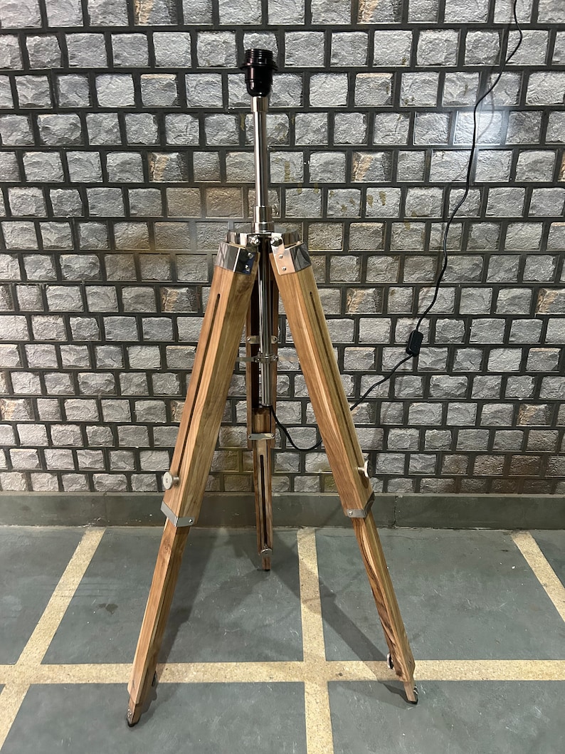 Puede incluir: Una l&aacute;mpara de pie de madera con un tr&iacute;pode de metal plateado y una pantalla negra. La l&aacute;mpara est&aacute; sobre un suelo de baldosas.