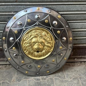 May include: A circular shield in silver with a gold lion's head centrepiece. The shield features a decorative border with rivets and gold detailing. The metal shield is likely for display or costume use. The diameter is approximately 76 cm.