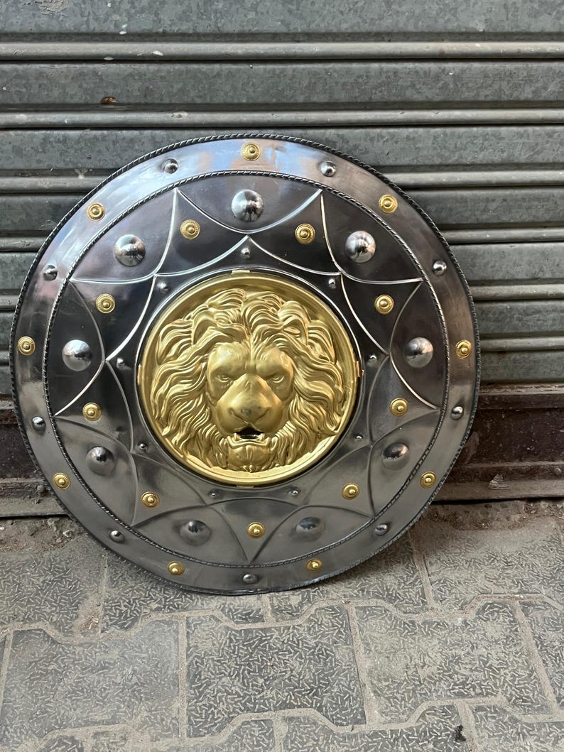 May include: A circular shield in silver with a gold lion's head centrepiece. The shield features a decorative border with rivets and gold detailing. The metal shield is likely for display or costume use. The diameter is approximately 76 cm.