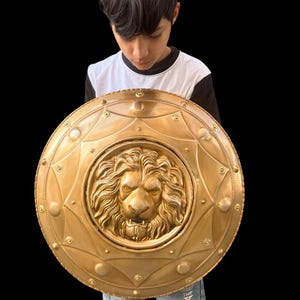 May include: A gold-coloured shield featuring a detailed lion's head at its centre. The circular shield has decorative rivets and a starburst pattern. The shield is held by a person wearing a black and white top and blue jeans.