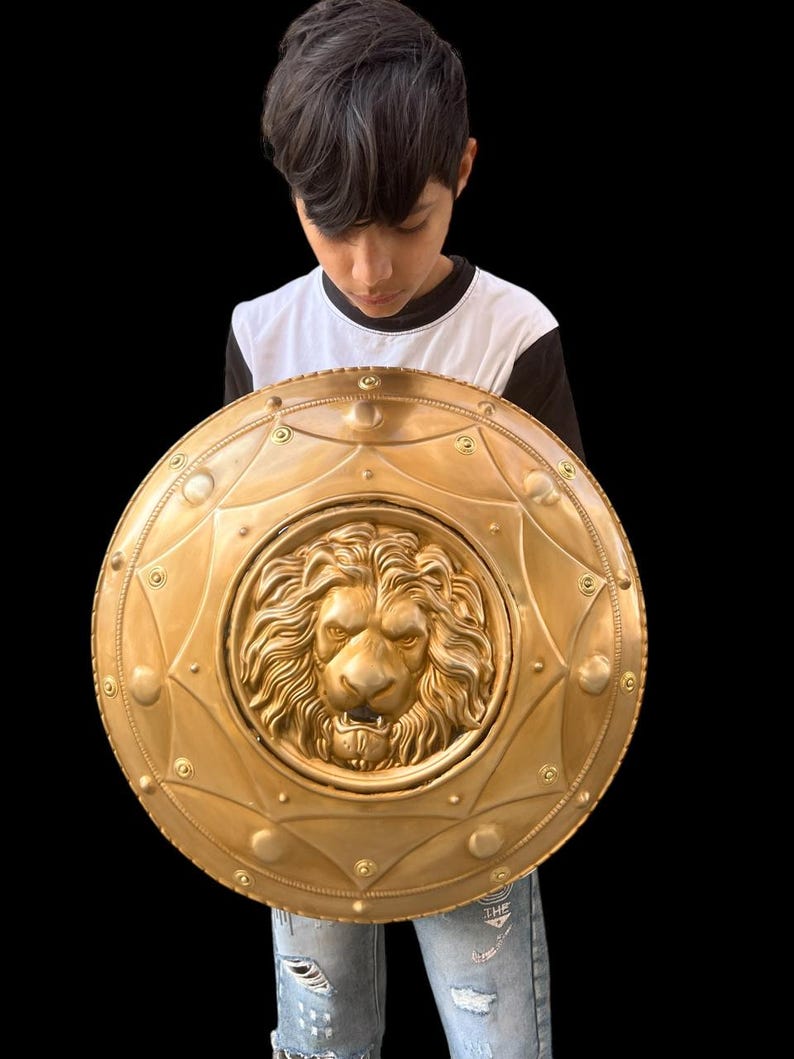 May include: A gold-coloured shield featuring a detailed lion's head at its centre. The circular shield has decorative rivets and a starburst pattern. The shield is held by a person wearing a black and white top and blue jeans.