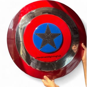 Puede incluir: Un escudo redondo con un anillo exterior rojo, un anillo central plateado y un anillo interior rojo. El centro presenta un c&iacute;rculo azul con una estrella negra. El texto "Handmadelightinghub" est&aacute; impreso en la parte inferior.