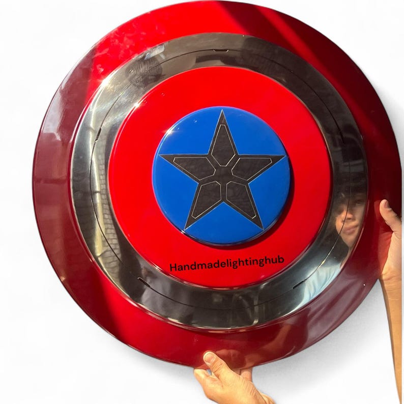 Puede incluir: Un escudo redondo con un anillo exterior rojo, un anillo central plateado y un anillo interior rojo. El centro presenta un c&iacute;rculo azul con una estrella negra. El texto "Handmadelightinghub" est&aacute; impreso en la parte inferior.