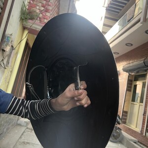 May include: A large, black, round shield with a curved handle. The shield is held by a person wearing a blue and white striped sleeve. The shield appears to be made of metal and has a leather strap.