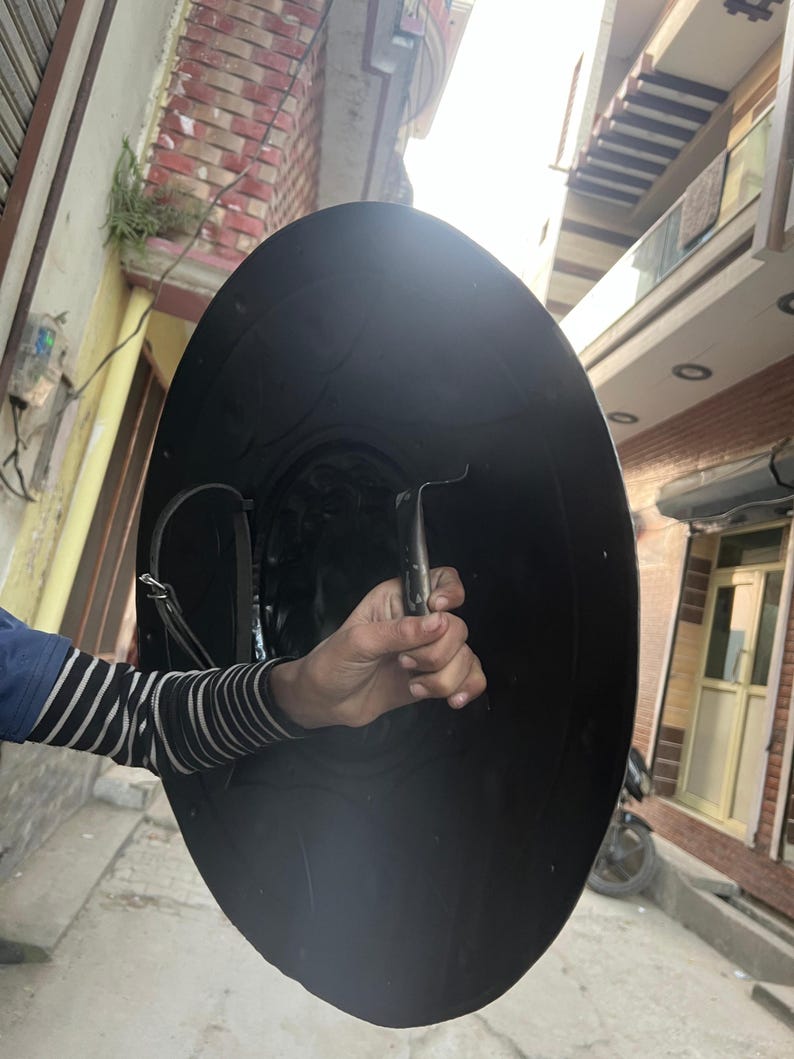May include: A large, black, round shield with a curved handle. The shield is held by a person wearing a blue and white striped sleeve. The shield appears to be made of metal and has a leather strap.