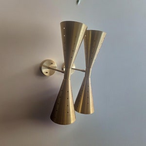Puede incluir: Un par de apliques de pared de color dorado con un diseño moderno. Cada aplique tiene dos brazos que se extienden desde una base central, y cada brazo tiene una pantalla cónica con un patrón perforado.