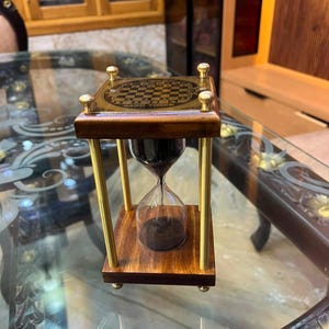 May include: A square hourglass featuring a dark wood base and top, four polished brass pillars, and a glass bulb filled with black sand. The top surface displays a gold-toned chessboard pattern with the text SAND TIMER printed in black.