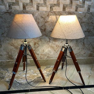 May include: Two tripod floor lamps with beige fabric lampshades. The lamps have wooden legs and silver accents.