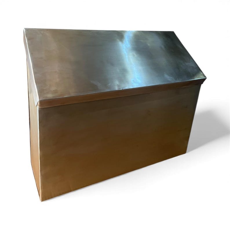 May include: A rectangular metal storage chest with a smooth, reflective silver-toned finish. The container features a slanted, hinged lid that slopes downward toward the front, creating a clean, angular silhouette with sharp, defined edges.