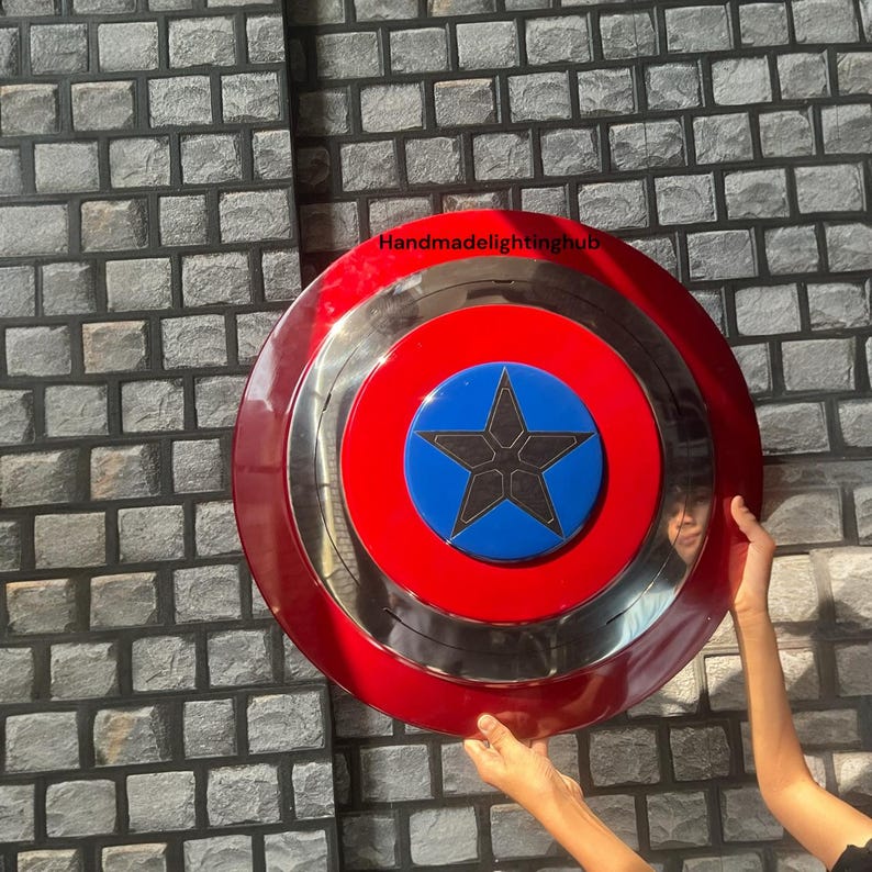 Puede incluir: Un escudo circular rojo, plateado y azul, con un emblema de estrella. El dise&ntilde;o incluye un anillo exterior rojo, un anillo central plateado y un centro azul con una estrella negra. El escudo est&aacute; sostenido por una persona.