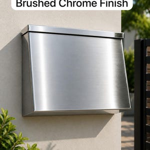 May include: A wall-mounted mailbox with a brushed chrome finish. The rectangular metal unit features a slanted front panel and a top lid. The text "Brushed Chrome Finish" is displayed in a white rounded rectangular box above the mailbox.