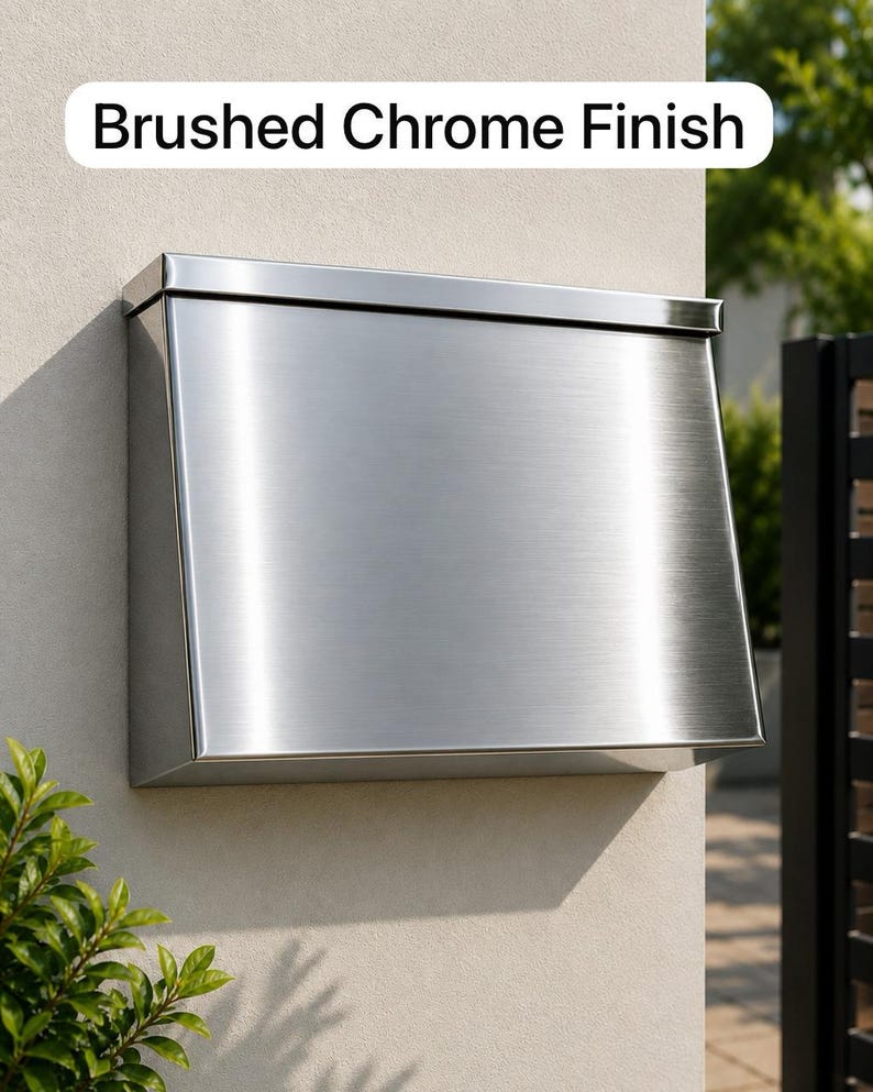 May include: A wall-mounted mailbox with a brushed chrome finish. The rectangular metal unit features a slanted front panel and a top lid. The text "Brushed Chrome Finish" is displayed in a white rounded rectangular box above the mailbox.