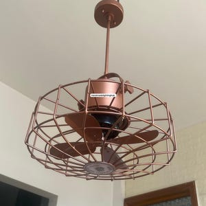 Può includere: Un ventilatore a soffitto color rame con un design a gabbia. Il ventilatore ha tre pale ed è sospeso dal soffitto da un'asta di metallo. Il ventilatore è etichettato "Handmadelightinghub".