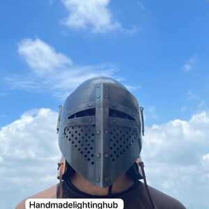 Pode incluir: Um close-up de uma pessoa usando um capacete de metal cinza escuro com uma viseira de malha e tiras de couro. O capacete tem uma superf&iacute;cie texturizada e &eacute; decorado com rebites. O fundo &eacute; um c&eacute;u azul com nuvens brancas. O texto "Handmadelightinghub" &eacute; vis&iacute;vel na parte inferior da imagem.