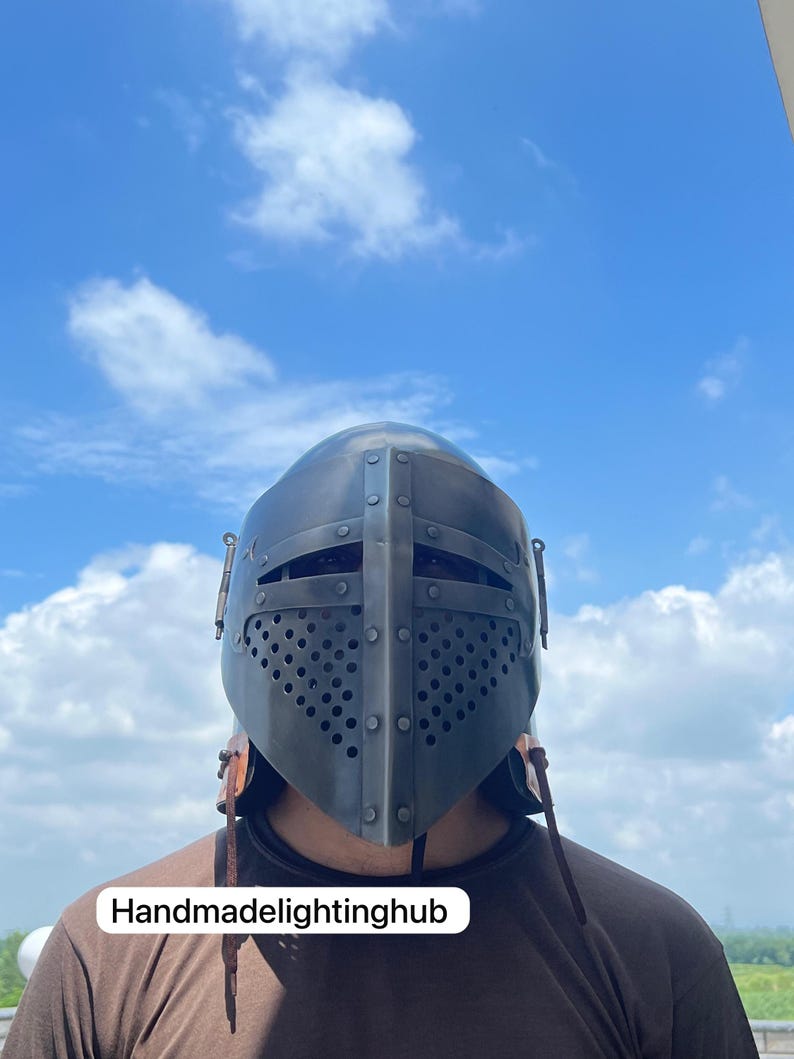 Pode incluir: Um close-up de uma pessoa usando um capacete de metal cinza escuro com uma viseira de malha e tiras de couro. O capacete tem uma superf&iacute;cie texturizada e &eacute; decorado com rebites. O fundo &eacute; um c&eacute;u azul com nuvens brancas. O texto "Handmadelightinghub" &eacute; vis&iacute;vel na parte inferior da imagem.
