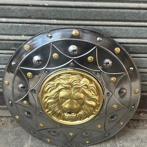 May include: A circular metal shield featuring a gold lion's head at its centre. The shield has a silver-grey finish with gold rivets and accents. The design is symmetrical, with a starburst pattern around the lion's head.