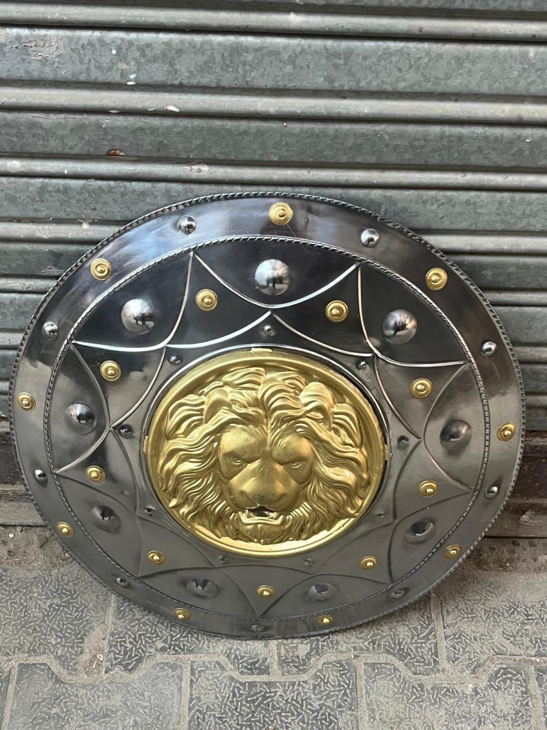 May include: A circular metal shield featuring a gold lion's head at its centre. The shield has a silver-grey finish with gold rivets and accents. The design is symmetrical, with a starburst pattern around the lion's head.