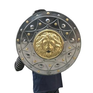 May include: A circular shield in silver with a gold lion's head emblem. The shield features a decorative border with rivets and gold detailing. The shield is held by a person wearing a dark blue top and jeans.