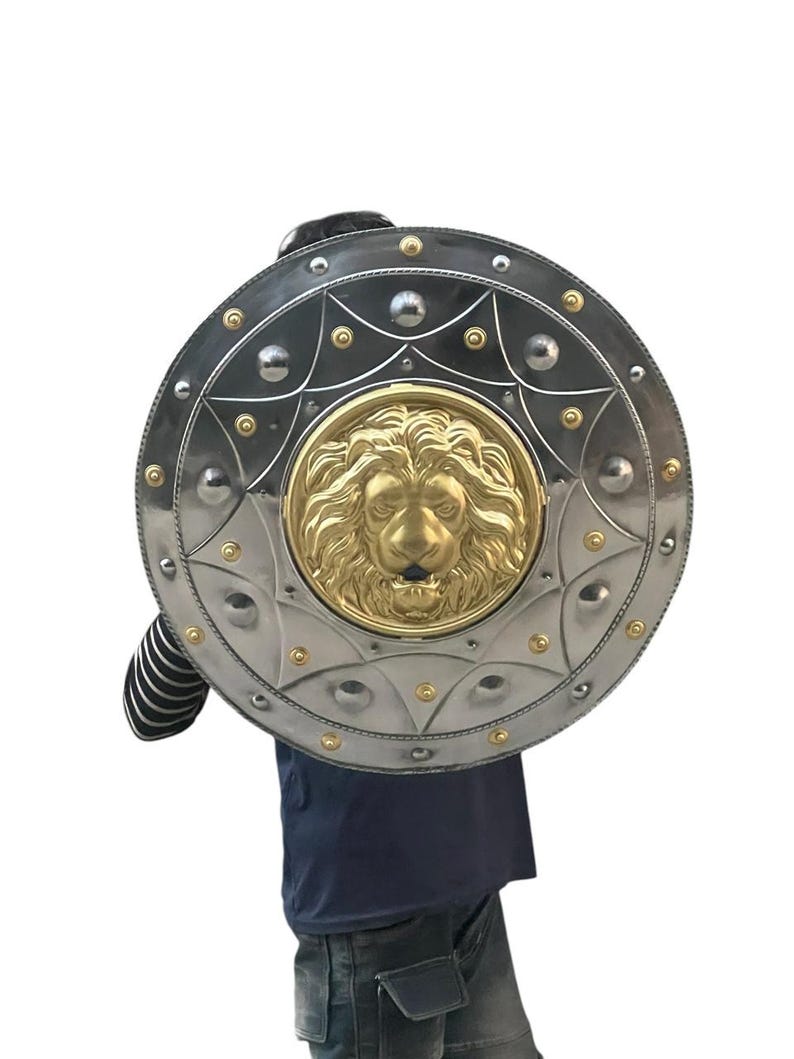 May include: A circular shield in silver with a gold lion's head emblem. The shield features a decorative border with rivets and gold detailing. The shield is held by a person wearing a dark blue top and jeans.