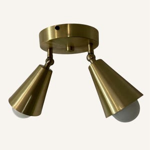 Puede incluir: Una lámpara de techo de dos luces de color latón. La lámpara tiene una base circular con dos luces ajustables en forma de cono. Cada luz tiene una bombilla blanca. La lámpara está diseñada para montarse en el techo.