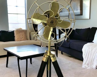 May include: A brass desk fan with a circular wire cage and four blades, mounted on a black wooden tripod stand. The center of the fan features a circular emblem with the text "MADE FOR ROYAL NAVY LONDON" and a small ship illustration in the middle.