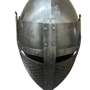 Pode incluir: Um capacete medieval com m&aacute;scara facial e viseira. O capacete &eacute; feito de metal e tem um acabamento cinza escuro. O capacete tem uma inscri&ccedil;&atilde;o "HELM" na parte superior e nas laterais.