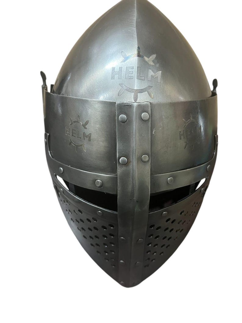 Pode incluir: Um capacete medieval com m&aacute;scara facial e viseira. O capacete &eacute; feito de metal e tem um acabamento cinza escuro. O capacete tem uma inscri&ccedil;&atilde;o "HELM" na parte superior e nas laterais.