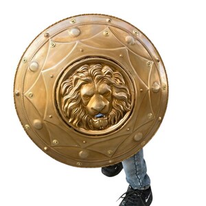 May include: A gold-coloured shield featuring a detailed lion's head at its centre. The shield is circular, adorned with decorative rivets and geometric patterns. The lion's face is intricately sculpted, displaying a fierce expression. The shield is held by a person wearing blue jeans and black trainers.