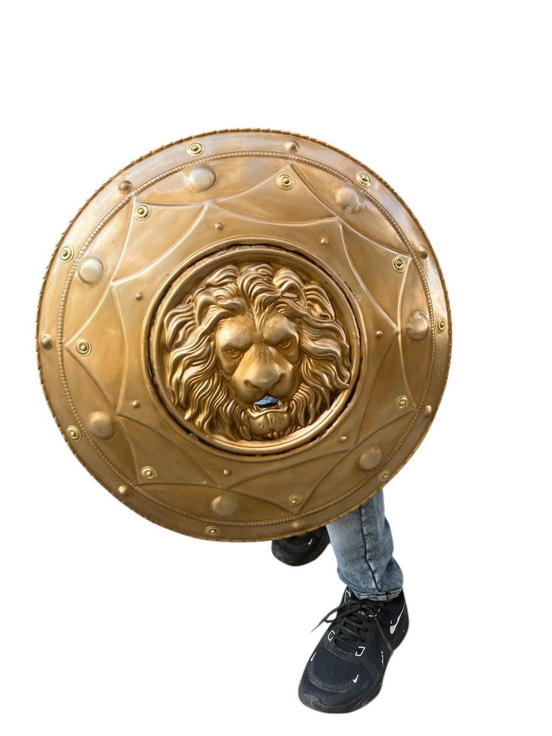 May include: A gold-coloured shield featuring a detailed lion's head at its centre. The shield is circular, adorned with decorative rivets and geometric patterns. The lion's face is intricately sculpted, displaying a fierce expression. The shield is held by a person wearing blue jeans and black trainers.