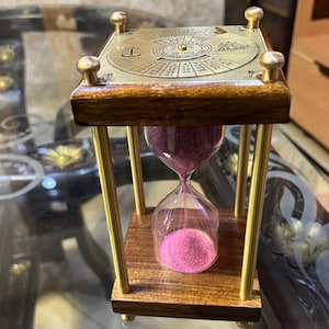 Puede incluir: Un reloj de arena con marco de madera y pilares de latón, con una placa superior que presenta un calendario grabado y un símbolo de ancla. El bulbo de vidrio contiene arena rosa vibrante que fluye de la cámara superior a la inferior.