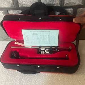 May include: A black carrying case with a red interior, containing a planimeter tool. The case has a zipper closure and a handle. The planimeter is used for measuring the area of irregular shapes. A table of standard settings in metric systems is visible, with the constant values 21 and 22.