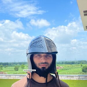 Pode incluir: Uma pessoa usando um capacete preto com um design de malha de metal e uma correia de couro. O capacete tem uma parte superior pontiaguda e uma parte traseira arredondada. A pessoa est&aacute; vestindo uma camisa marrom e tem barba.