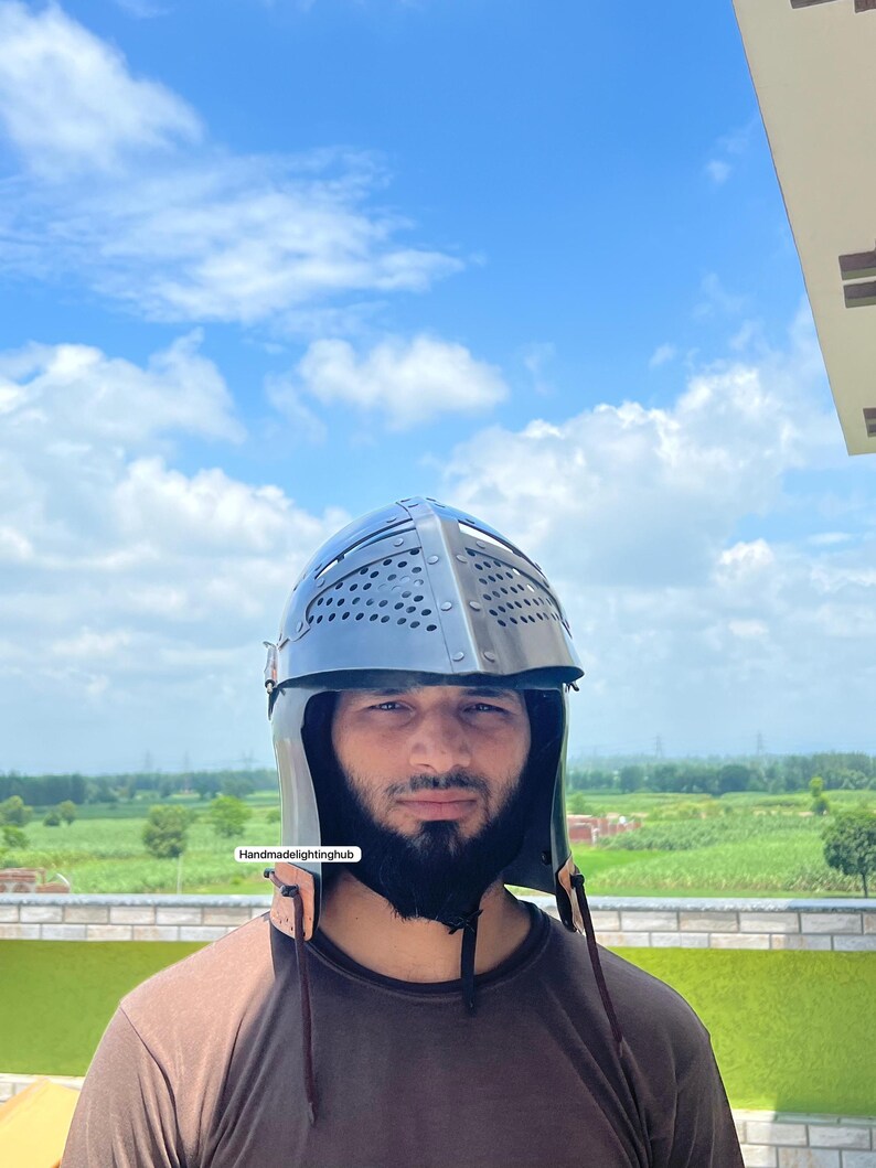 Pode incluir: Uma pessoa usando um capacete preto com um design de malha de metal e uma correia de couro. O capacete tem uma parte superior pontiaguda e uma parte traseira arredondada. A pessoa est&aacute; vestindo uma camisa marrom e tem barba.