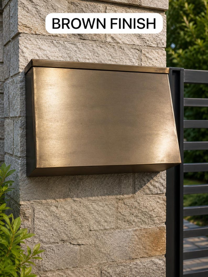 May include: A rectangular wall-mounted mailbox with a smooth brown finish. The mailbox features a slanted front panel and a top lid. A white rectangular label with the black text BROWN FINISH is positioned above the mailbox on the stone wall.