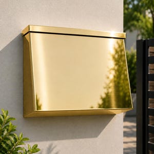 May include: A rectangular wall-mounted mailbox with a polished gold-tone metallic finish. The mailbox features a slanted front panel that reflects the surrounding environment and a flat top lid, securely attached to a light-colored exterior wall surface.
