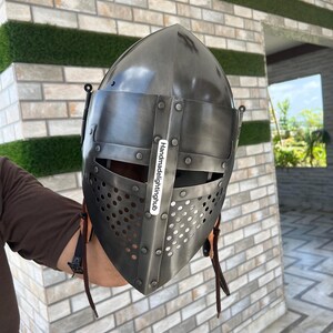 Pode incluir: Um capacete medieval com uma parte superior pontiaguda e uma m&aacute;scara facial com um design perfurado. O capacete &eacute; feito de metal e tem uma correia de couro marrom.