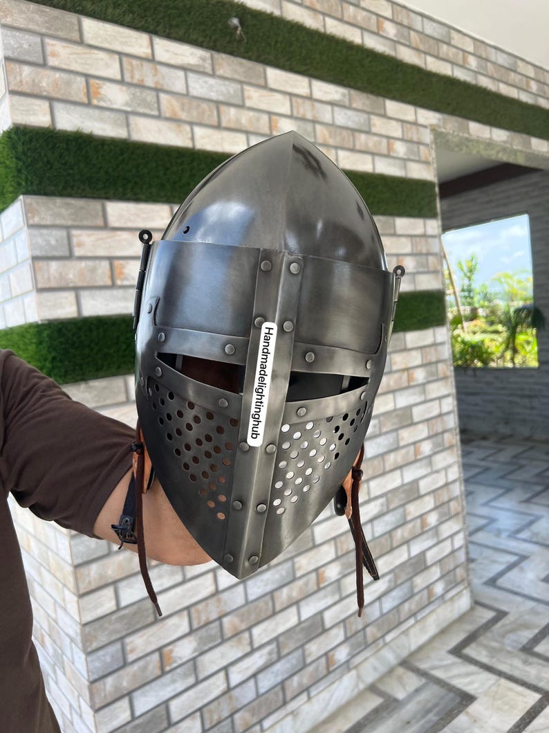 Pode incluir: Um capacete medieval com uma parte superior pontiaguda e uma m&aacute;scara facial com um design perfurado. O capacete &eacute; feito de metal e tem uma correia de couro marrom.