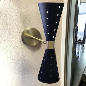 May include: A black wall sconce with two cone-shaped shades featuring a perforated design. The sconce has a brass base and arm.