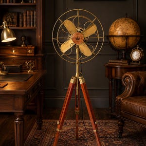May include: A floor-standing electric fan featuring four brass-coloured blades and a circular wire cage. The fan is mounted on a three-legged wooden tripod base with brass hardware. A circular emblem with a crown icon and text is centred on the fan.