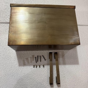 May include: A rectangular metal mailbox with a brown finish, accompanied by two mounting brackets, four screws, and four small fasteners. The text "BROWN FINISH" is displayed above the mailbox, indicating the color of the metal surface.