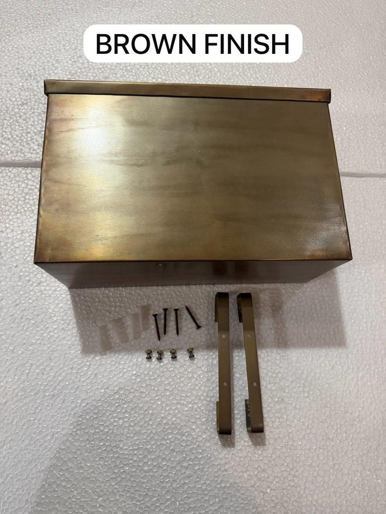 May include: A rectangular metal mailbox with a brown finish, accompanied by two mounting brackets, four screws, and four small fasteners. The text "BROWN FINISH" is displayed above the mailbox, indicating the color of the metal surface.